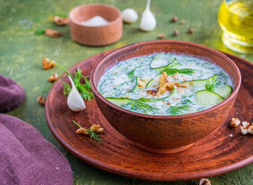 Traditional Bulgarian cold soup tarator with cucumber, herbs and walnuts in a brown clay bowl on a green concrete background. Cold soup recipes.