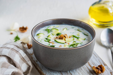 Traditional Bulgarian cold soup tarator with cucumber, herbs and walnuts in a gray bowl on a light concrete background.