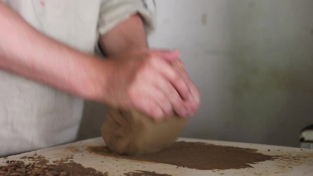 a male potter of Caucasian nationality in a working uniform kneads clay on a rbotemstol in his workshop, close-up. Pottery preparation for sculpting