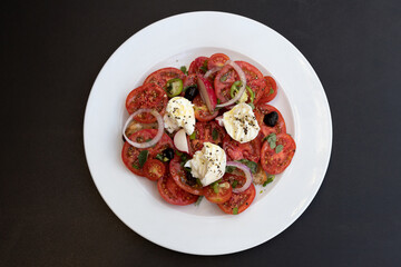 Italian caprese salad with chopped tomatoes, mozzarella, basil, oregano, thassos, onions, radishes, chili peppers and olive oil on a white plate.