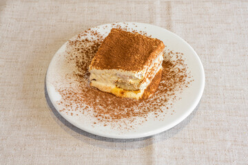 Piece of tiramisu cake with its typical layers sprinkled with cocoa on a round white plate