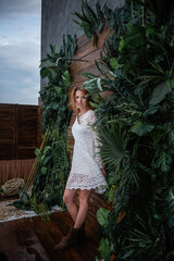 A young woman in a white openwork dress, brown boots stands against a wooden background wall with tropical green plants: monstera, ficus, palm. Fashion girl on a journey, summer interior decoration