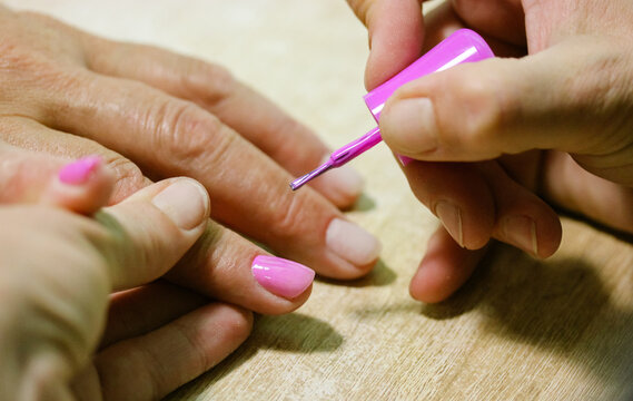 A manicurist applies pink gel shellac nail polish with a brush on a client's nails in a beauty salon. Spa treatments. Women's hands and fingers at the wooden table up close. Female nail care process.