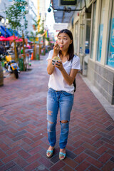 Happy Young Asian Woman Wearing Distressed Jeans and T-Shirt Eating an Ice Cream Sandwich in a Downtown Urban Area