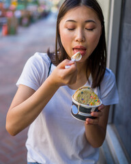 Happy Young Asian Woman Wearing Distressed Jeans and T-Shirt Eating an Ice Cream Sandwich in a Downtown Urban Area
