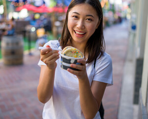 Happy Young Asian Woman Wearing Distressed Jeans and T-Shirt Eating an Ice Cream Sandwich in a Downtown Urban Area