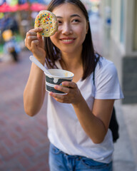 Happy Young Asian Woman Wearing Distressed Jeans and T-Shirt Eating an Ice Cream Sandwich in a Downtown Urban Area