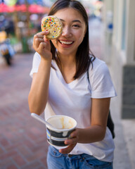Happy Young Asian Woman Wearing Distressed Jeans and T-Shirt Eating an Ice Cream Sandwich in a Downtown Urban Area