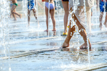 A barefoot young woman in a white dress escapes the heat in splashes of water in a city fountain.
