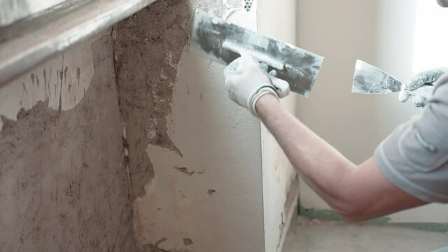 Repairman in working clothes spackling ragged cement wall under the window using metal trowel and white plaster paste, builder leveling wall surface with spatula and putty stucco. Walls restoration