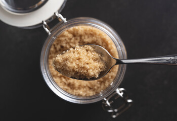 Brown sugar and a spoon in a glass container set against a black stone background.