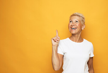 Senior grey-haired woman wearing white t-shirt pointing finger up with successful idea over yellow...
