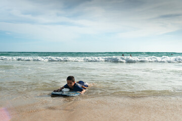 Fototapeta premium The waves swept the surf boy toward the shore..boy surfing on white waves..Paradise beach blue sea, and clear sand landscape background..smiling face of happy boy surfing concept.