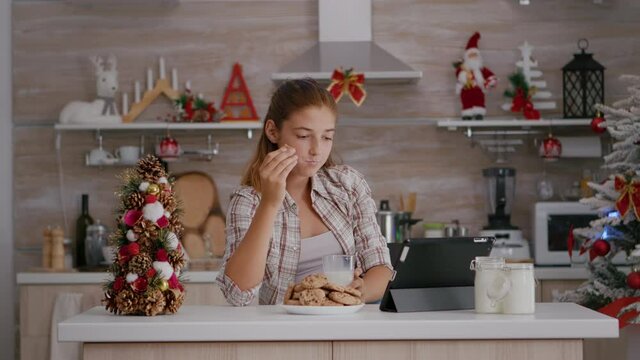 Portrait of children eating baked delicious cookies drinking milk while watching online xmas video on tablet computer in decorated kitchen. Caucasian girl celebrating christmas enjoying winter holiday