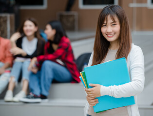 Close up young attractive female college students holding document files smiling at camera with blurry university campus and other students. Outdoor. Concept for education, college students life