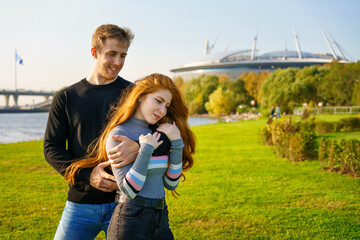 beautiful young couple, man and woman with long red hair, stand in an embrace in city park against the backdrop of modern buildings and green grass. Happy and romantic guy and girl on sunny day