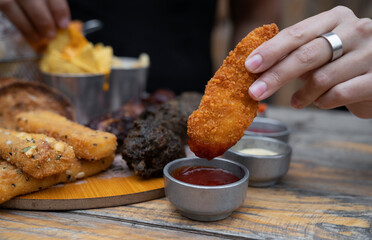 Couple sharing a meal. Closeup view of a caucasian woman's hand dipping a fried chicken stick in ketchup. In the background, mozzarella sticks and a male hand holding a nacho with cheddar cheese. 