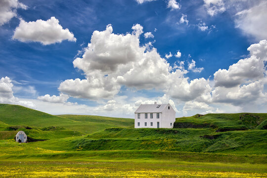 A square white house on a hill among green fields. In summer there are many yellow wildflowers, isolated houses and striking nature in the Icelandic countryside.