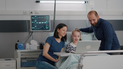 Obraz premium Family sitting beside little daughter greeting friends during online videocall conference meeting in hospital ward. Chid patient resting in bed after suffering examination surgery