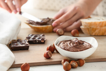 Woman spreading tasty chocolate paste with hazelnuts on bread in kitchen