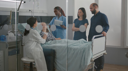 Pediatric doctor examining breathing sickness symptoms while african american nurse checking IV drip working in hospital ward. Sick little kid resting in bed recoverying after disease surgery