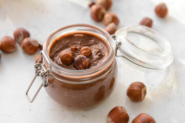 Jar with tasty chocolate paste and hazelnuts on light background