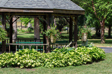 Close up view of large wooden gazebo landscaped with beautiful hostas and hanging planters, in a public park on a sunny summer day