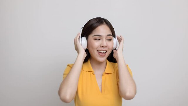 Cheerful asian female teenager listen to the music with white headphone dancing on isolated. Beautiful young woman in yellow shirt hand touch a wireless headphone having fun with the music in  room.