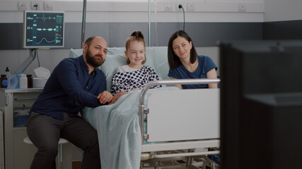 Hospitalized sick little patient lying in bed with family watching entertainment movie on television during medical recovery examination in hospital ward. Kid waiting for disease treatment