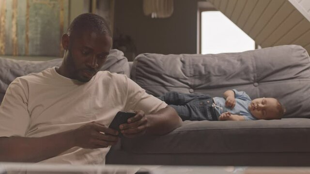 Waist-up of caring African man sitting by couch at home, using smartphone for taking picture of cute Mixed-Race baby
