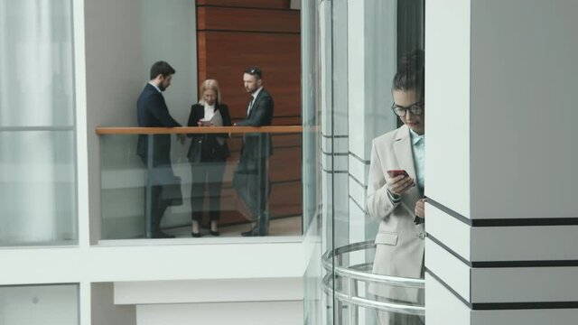 Tilt-up slowmo shot of young brunette female lawyer going up in elevator while team of her colleagues having conversation standing by railing in modern office