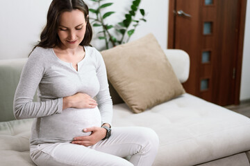beautiful pregnant woman in grey jacket sitting on couch touching pregnant belly, happy waiting