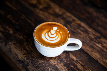 white cup of cappuccino with latte art on saucer with spoon on dark wooden background.