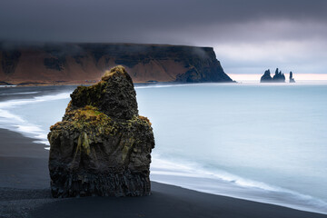Arnardrangur basalt rock at Kirkjufjara beach looking to scenic Reynisfjara black sand beach and Reynisdrangar during sunrise on a stormy cloudy morning in Iceland