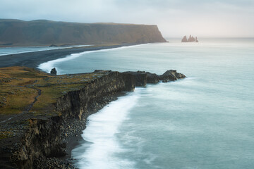 Dramatic stormy sunrise with clouds, fog and sunshine at scenic Kirkjufjara cliff looking to Reynisfjara black sand beach and Reynisdrangar in Iceland