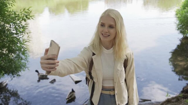 A young blonde woman with a backpack takes pictures of herself on the phone in the park against the backdrop of a pond. Ducks and pigeons fly behind her. Travel and adventure concept. Slow motion, HD.