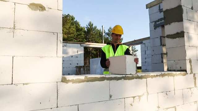 Construction worker at construction site puts the wall of a house made of aerated concrete blocks. Protective clothing - hardhat and a vest, the construction process