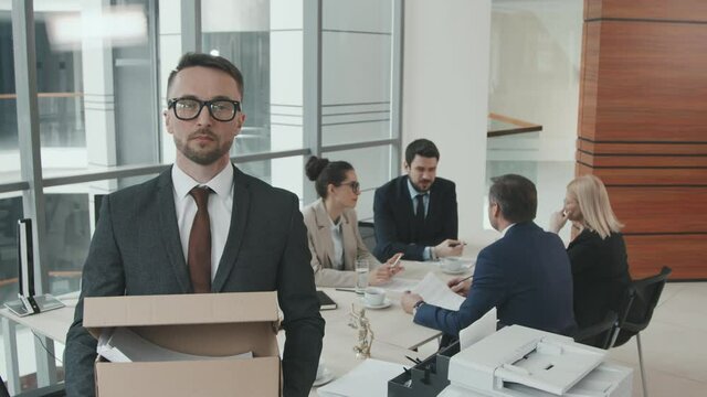 PAN medium slowmo portrait of young male lawyer in suit and eyeglasses holding cardboard box of legal documents looking at camera while his colleagues discussing case in background