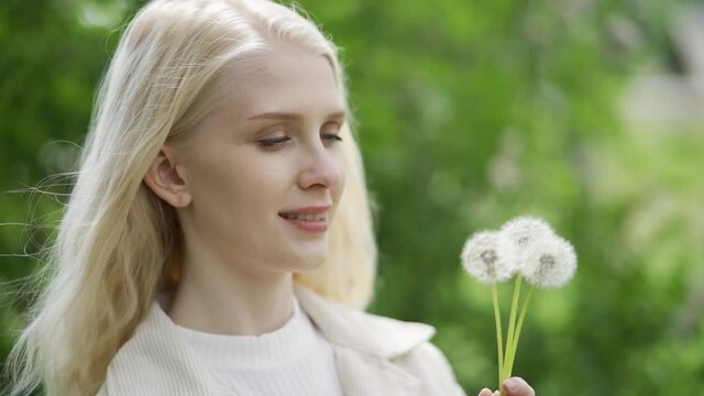 A romantic woman with curls fervently blows on dandelions. Naive young blond woman. Rest in the country. Slow motion, close-up, HD.