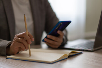 Female hand works in the phone near the laptop and notepad. Married woman in jacket working at home office. Horizontal view.