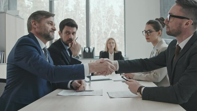 Medium slowmo of two successful businessmen and their attorneys sitting at negotiating table in modern office room shaking hands after making deal