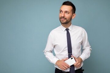 Handsome thoughtful good looking brunet unshaven man with beard wearing casual white shirt and tie isolated on blue background with empty space holding in hand mobile phone looking to the side