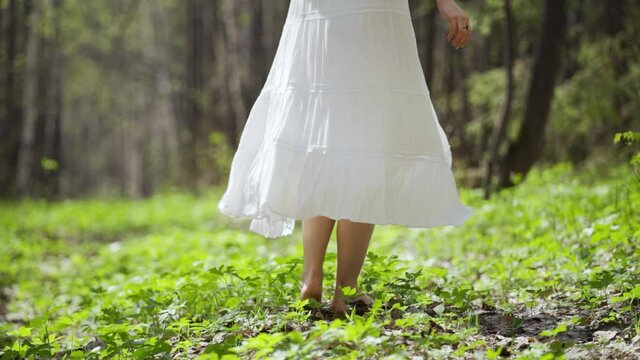 A romantic woman in a light white dress whirls barefoot in the forest. The joy of being in nature. Warm sunny summer day. Slow motion, close-up, HD.