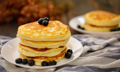 Stack of delicious pancakes sweet food with blueberries and blur dry leaves, plaid fabric in white plate on wooden background.