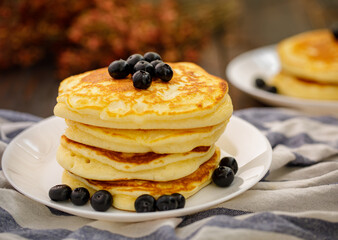 Stack of delicious pancakes sweet food with blueberries and blur dry leaves, plaid fabric in white plate on wooden background.