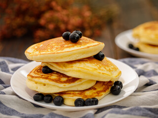 Stack of delicious pancakes sweet food with blueberries and blur dry leaves, plaid fabric in white plate on wooden background.