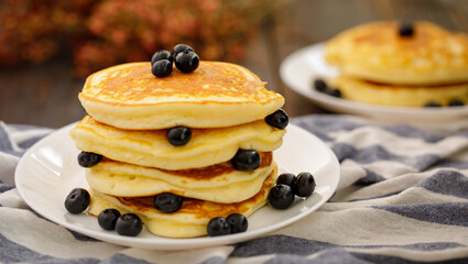 Stack of delicious pancakes sweet food with blueberries and blur dry leaves, plaid fabric in white plate on wooden background.