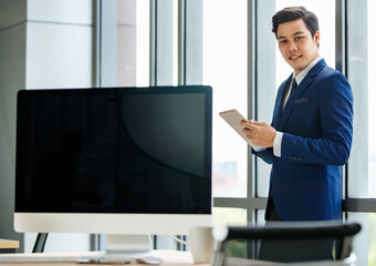 Portrait of young and good lookiing Asian businessman is blue suit standing and holding tablet in modern office with screen of desktop computer in blur foreground