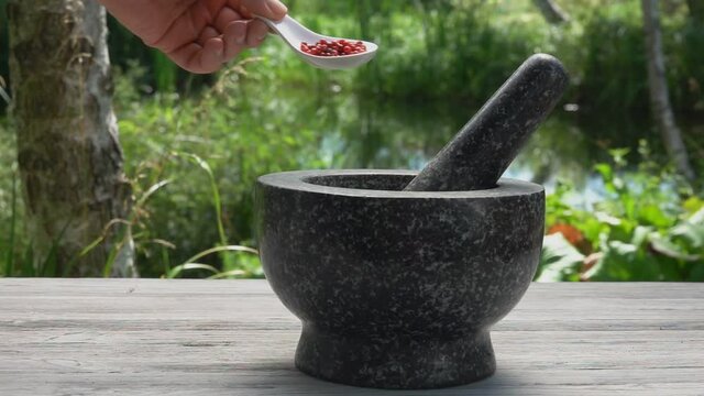 Hand is putting the rose peppercorns into a grey mortar in slow motion outdoors