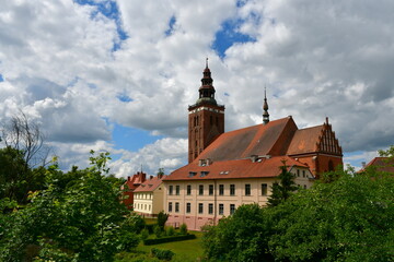An old town or village with a big church with a tall tower made out of red brick and metal in the middle seen from a tall hill full of stones, lamps and covered with a wall made out of red brick